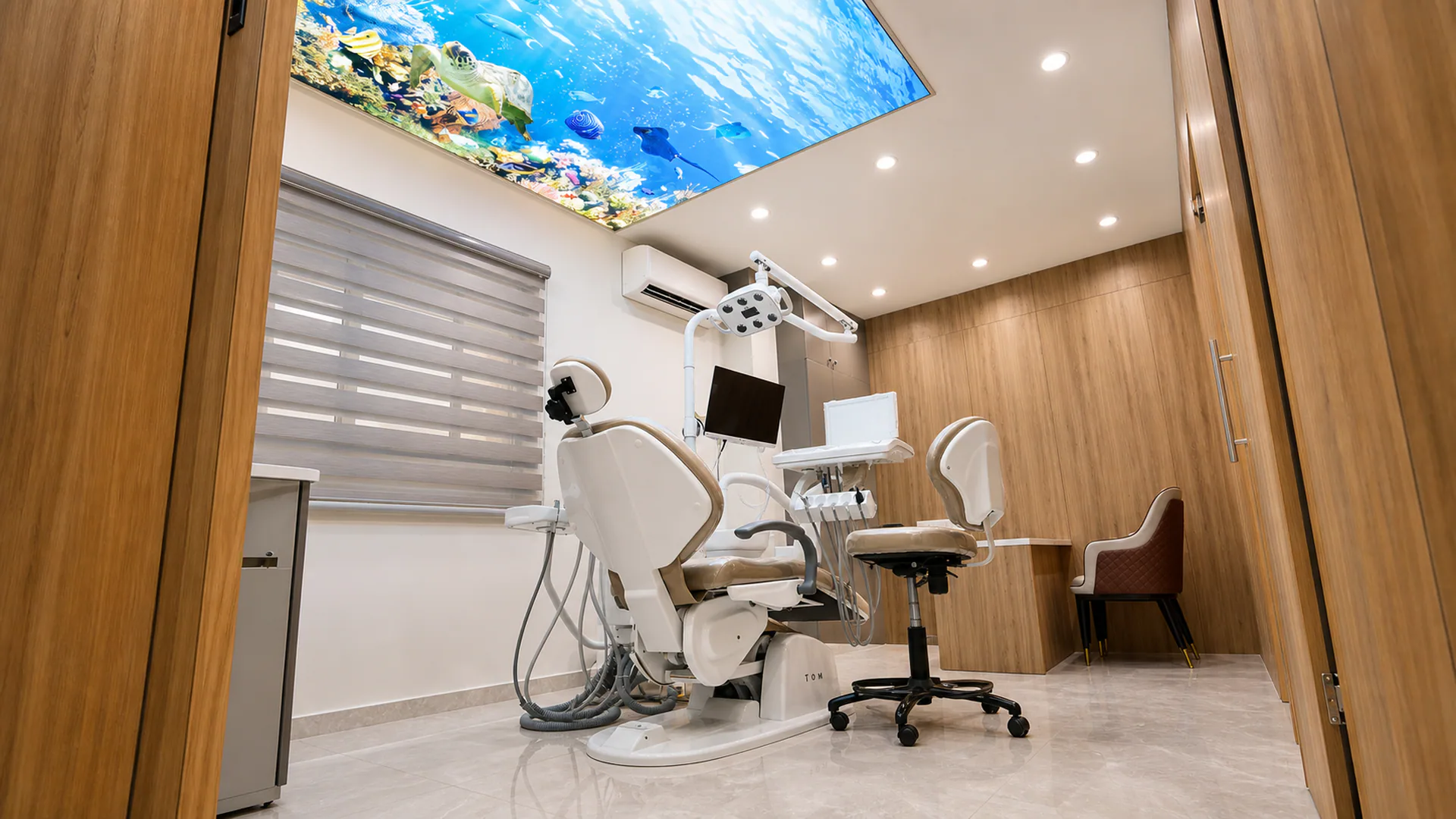 Dental treatment room with chair, equipment, wood-finished walls and an ocean-themed ceiling panel.