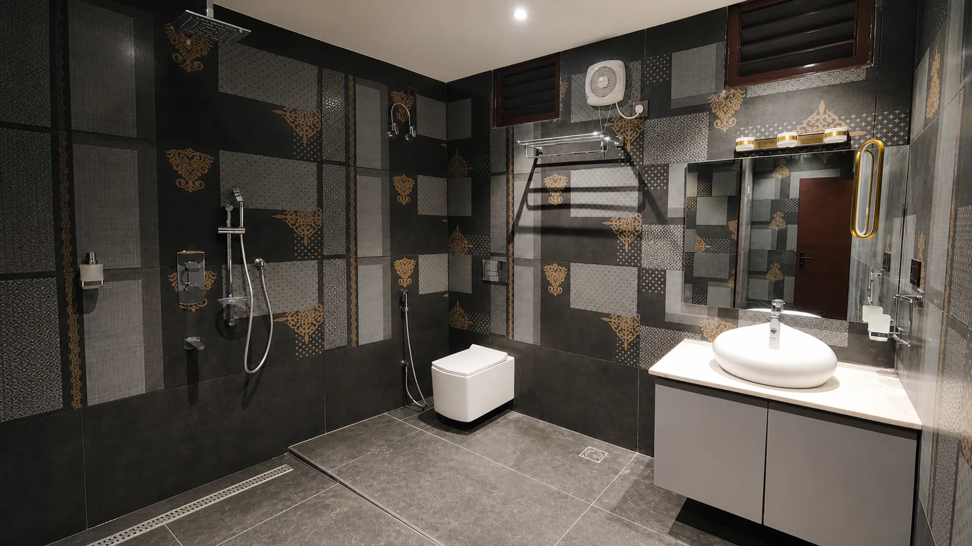 Bathroom with dark patterned wall tiles, shower fittings, toilet and wall-mounted basin.